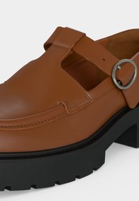 Καφέ δερμάτινο loafer με κοψίματα, ασημένιο στρογγυλό πόρπη και χοντρή μαύρη σόλα με αυλακώσεις.