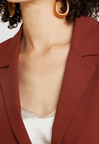 Blazer couleur rouille avec une texture lisse, doté de revers. En dessous, un haut en satin léger avec un léger éclat. Grandes boucles d'oreilles en forme de cercle orange.