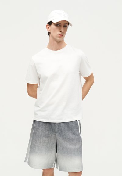 Νέος άνδρας με άσπρο καπέλο και απλό λευκό t-shirt, όρθιος με τα χέρια πίσω από την πλάτη, φορώντας γκρι σορτς με διαβάθμιση χρωμάτων, σε ένα φωτεινό φόντο.