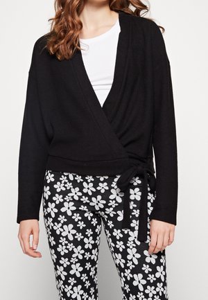 Vrouw draagt een zwarte wikkelsweater over een wit shirt met zwarte broek met een wit bloemmotief, staand tegen een effen achtergrond.