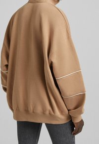 Sweatshirt beige surdimensionné avec col haut, poignets côtelés et rayures blanches contrastantes sur les manches. Texture douce, coupe décontractée et silhouette relaxée.