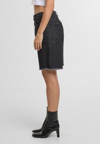 Mujer vestida con una falda de mezclilla negra de cintura alta con dobladillo deshilachado y botaschel de tacón negras, de pie contra un fondo liso.