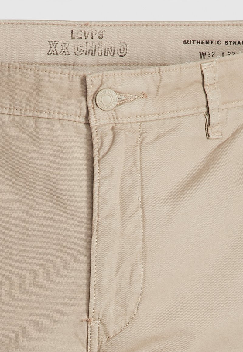 Pantalons chinos Levi's XX beiges avec une texture lisse, fermeture à bouton et détails cousus. L'étiquette indique "Authentic Straight" en marron.