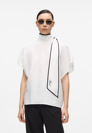 Žena s uhladenými vlasmi má na sebe čierne slnečné okuliare, voľnú bielu tuniku s vysokým golierom, čierne nohavice a biely šál s čiernym lemom a podpisom.