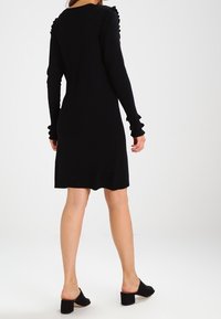 Svart klänning med långa ärmar och volanger på axlar och manschetter, tillverkad av ett mjukt tyg, med en knälång silhuett.