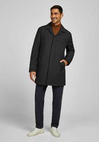 Zwarte jas met een Engelse kraag, lange mouwen en zijzakken. Draagt over een bruine trui en gecombineerd met marineblauwe broek en witte sneakers.