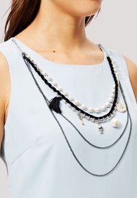 Collarín en capas que presenta cadenas negras, perlas blancas, acentos plateados y colgantes que incluyen un cráneo y una moneda, usado sobre una blusa azul clara.