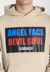 Sudadera beige con un gráfico frontal que presenta "ANGEL FACE" en azul, "DEVIL SOUL" en rojo y "KAVANAGH" en blanco, sobre un fondo negro.