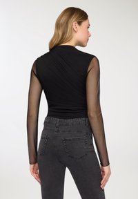 Top nero a maniche lunghe con maniche in rete trasparente, caratterizzato da un design drappeggiato sulla schiena. Abbinato a jeans grigio scuro a vita alta. Texture liscia.