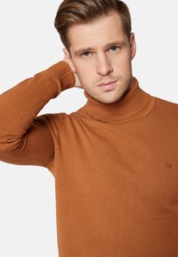 Brązowy sweter typu golf z prążkowanym wzorem, dopasowanym krojem i małym czerwonym logiem na lewej piersi. Miękka tkanina, długie rękawy.