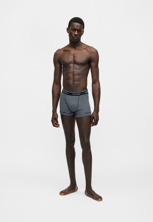 Αδύνατος σκουρόχρωμος άνδρας που στέκεται ξυπόλητος πάνω σε απλό φόντο, φορά μπλε και λευκά ριγέ εσώρουχα Calvin Klein.