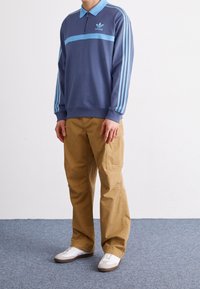 Modrý fleecový svetr s límečkem, předním zipem a světle modrými pruhy na rukávech, kombinovaný s béžovými cargo kalhotami a bílými teniskami.