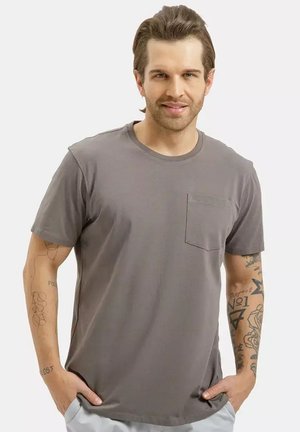 Mężczyzna z ułożonymi włosami i tatuażami na obu ramionach, ubrany w szarą koszulkę z krótkim rękawem i kieszenią na piersi, z rękami w kieszeniach jasnych spodni.