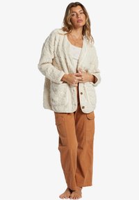 Cardigan in tessuto crema con un morbido motivo trapuntato, tasche frontali e chiusura con bottoni, abbinato a pantaloni a gamba larga color ruggine.