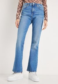 Vaqueros de mezclilla azul claro con diseño acampanado, cintura alta y un sutil desgastado. Combinados con zapatillas blancas para un contraste.