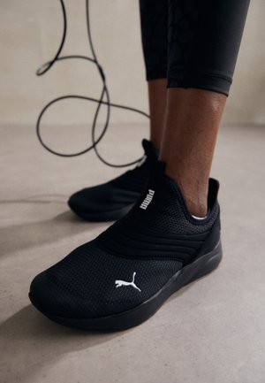 Gros plan sur une personne portant des baskets Puma noires et des leggings noirs, tenant une corde à sauter avec une corde bouclée sur un sol lisse.