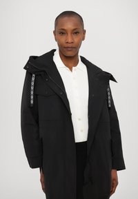 Parka noire imperméable avec capuche, quincaillerie argentée, poches avant et cordons à motifs contrastants. Superposé sur une chemise blanche à boutons.