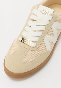 Sneaker beige con superiore in rete testurizzata e camoscio, dettagli bianchi, lacci spessi e placca dorata con la scritta "STEVE MADDEN". Sole in gomma.