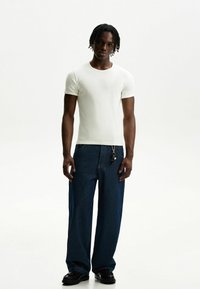 White short-sleeve t-shirt paired with loose-fitting dark blue denim pants, featuring a keychain attached to the waistband and black shoes.