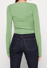 Top verde a maniche lunghe con un leggero luccichio, design aderente e scollo a barca, abbinato a jeans scuri con dettagli nella cucitura delle tasche.