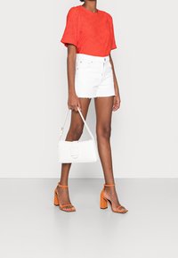 Blouse rouge à manches courtes avec détail en œillets, shorts en denim blanc déchiré, sac à main blanc avec boucle, et sandales à talons orange.