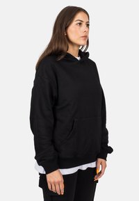 Sudadera negra de tejido suave, con un bolsillo delantero, capucha ajustable con cordón y puños acanalados. Llevada sobre una camiseta blanca.
