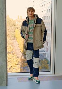 Beige og marineblå hettejakke over stripete t-skjorte, kombinert med slitte blå jeans med kremfargede paneler. Grønne og hvite joggesko.