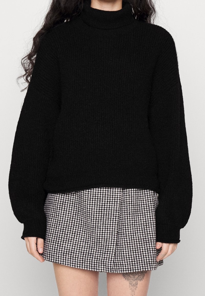 Femme portant un pull en maille noir et une jupe à carreaux noir et blanc, montrant un tatouage sur sa cuisse gauche supérieure.