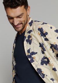 Korte mouwen, crème shirt met donkere bloemenprint en blauwe accenten; gelaagd over een donker marineblauw ronde hals top. Glad stoftextuur.