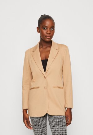 Kvinde med mørk hud og trukket hår tilbage iført en beige blazer over en sort top og sort-hvide mønstrede bukser, stående mod en hvid baggrund.