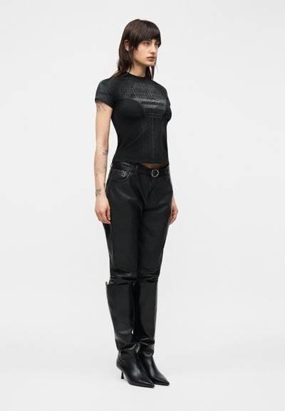 Γυναίκα με σκουρόχρωμα μαλλιά, που φοράει εφαρμοστή μαύρη μπλούζα με στάμπα, μαύρο δερμάτινο παντελόνι, μαύρη ζώνη και μαύρα μπότες με τακούνι μέχρι το γόνατο, σε λευκό φόντο.