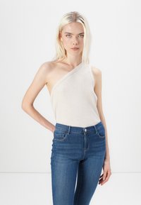 Top sin mangas de canalé beige con un tirante, combinado con jeans ajustados de color azul oscuro. Ambas prendas tienen texturas suaves y diseños ajustados.