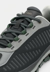 Szürke és fekete sportcipő texturált felsőrésszel, zöld és szürke fűzőkkel, valamint feltűnő dizájnvonalakkal az oldalon. Párnázott nyelvvel rendelkezik.