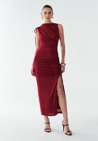 Vīna krāsas gara kleita ar asimetrisku plecu dizainu, kroku tekstūru un augšstilba augstuma šķēlumu, kombinēta ar melniem siksnainiem augstpapēžu apaviem.