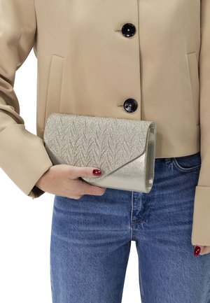 Clutch metallica oro con motivo fogliare in rilievo, design a patta triangolare, tenuta in mano. Abbinata a una giacca beige e jeans di denim.