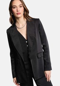 Blazer de satén negro con solapa de muesca, cierre de un botón y dos bolsillos delanteros. Llevado sobre una blusa negra con botones decorativos.