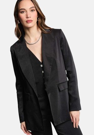 Blazer en satin noir avec un col cranté, fermeture à un bouton et deux poches avant. Porté par-dessus un haut noir avec des boutons décoratifs.