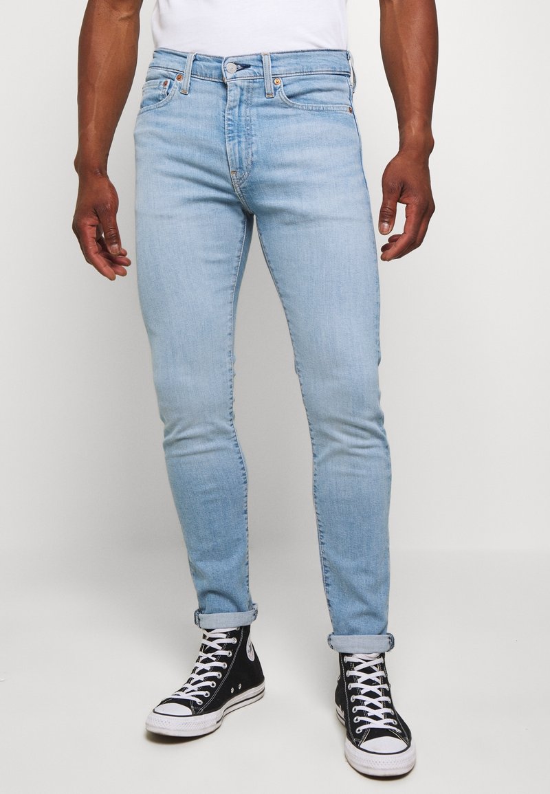 Homme portant un jean skinny bleu clair retroussé aux chevilles et des baskets montantes noires avec des lacets blancs sur fond blanc.