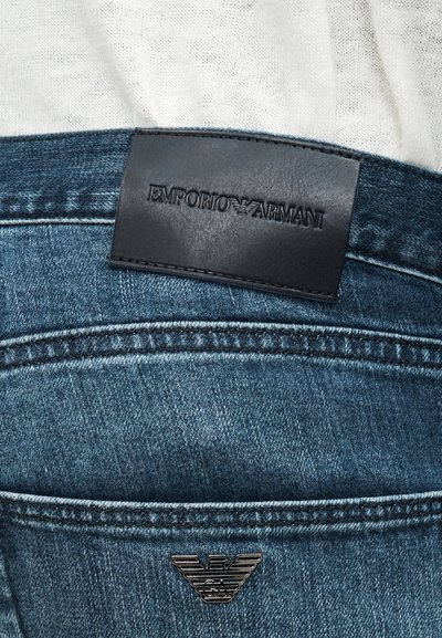 Κοντινή λήψη μπλε τζιν με μαύρη δερμάτινη ετικέτα Emporio Armani και μεταλλικό λογότυπο αετού στη ζώνη πίσω μέρος.