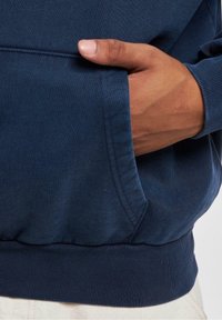 Granatowa bluza z teksturowanej tkaniny, z przednią kieszenią z prążkowanym wykończeniem i luźnym krojem. Ręka spoczywa w kieszeni.