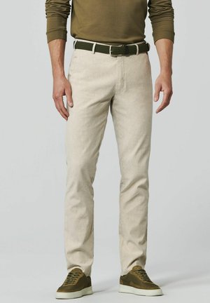 Mann trägt beige Hosen, olivgrünen Gürtel, olivgrünes Langarmshirt und olivgrüne Turnschuhe, steht vor einem einfarbigen Hintergrund.