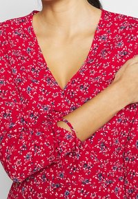 Rotes Kleid mit Floralprint, V-Ausschnitt und langen Ärmeln. Der Stoff wirkt leicht und hat eine weiche Textur. Die Ärmelbündchen sind mit Bindebändern versehen.