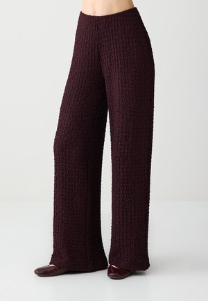 Szerokie spodnie z teksturowanego ciemnego burgundowego materiału noszone z dopasowanymi płaskimi butami, pokazane od talii do stóp na jednolitym jasnym tle.