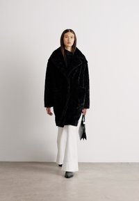 Palton din blană artificială neagră, cu un guler lat, design căptușit dublu, două buzunare frontale, purtat peste un top negru și pantaloni evazați albi.
