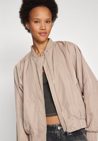 Veste bomber beige clair avec fermeture éclair à l'avant, col côtelé et poignets élastiques. Tissu lisse et légèrement brillant avec une coupe décontractée.