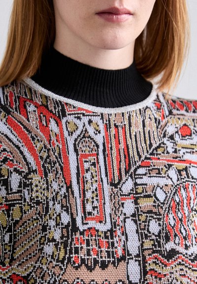 Pull en tricot avec un col montant noir côtelé. Il présente un motif géométrique coloré en rouge, or et noir sur un fond crème.