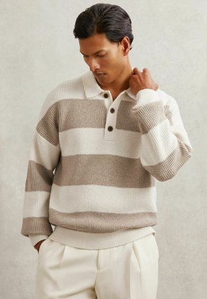Mężczyzna o ciemnych włosach, noszący beżowy i biały pasiasty sweter w wafle z guzikami, dotykający szyi, stojący na neutralnym tle.