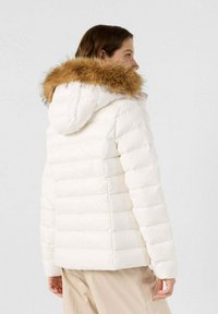 Chaqueta puffer blanca con un diseño texturizado y acolchado. Presenta una capucha con un borde de pelo sintético marrón y mangas largas.