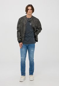 Giacca di pelle nera con accenti a coste sulle spalle, chiusura zip, abbinata a una t-shirt grigia scura, jeans strappati blu e sneakers bianche.