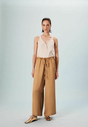 Haut sans manches crème avec un détail en trou de serrure, associé à un pantalon large beige avec une taille élastique et un cordon de serrage. Chaussures métalliques.